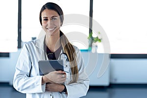 Happy young female doctor smiling and looking at camera while digital tablet and standing in the consultation
