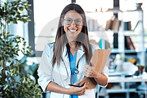 Happy young female doctor smiling and looking at camera while holding a clipboard and standing in the consultation