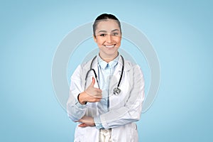 Happy young female doctor with stethoscope around her neck giving thumbs up and smiling, blue backdrop