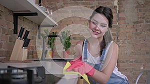 Happy young female housewife in rubber gloves for cleaning wipe dusty furniture on kitchen