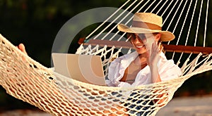 Happy young female wearing straw hat waving while having video call on digital laptop, relaxing in the hammock on tropical beach