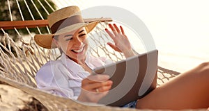 Happy young female wearing straw hat waving while having video call on digital tablet, relaxing in the hammock on tropical beach