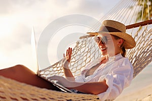 Happy young female wearing straw hat waving while having video call on laptop, relaxing in the hammock on tropical beach