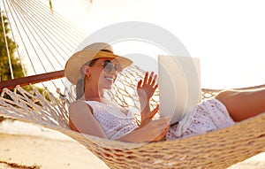 Happy young female wearing straw hat waving while having video call on laptop, relaxing in the hammock on tropical beach
