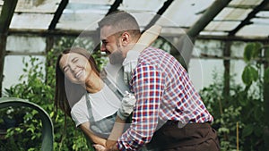 Happy young florist couple in apron have fun while working in greenhouse. Laughing woman spray water in husband face