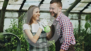 Happy young florist couple in apron have fun while working in greenhouse. Laughing woman spray water in husband face