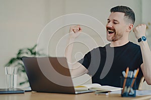 Happy young freelancer showing winning gesture while sitting at his workplace at home