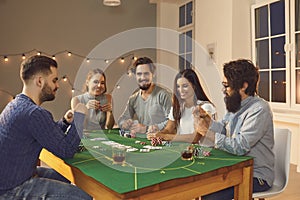 Happy young friends playing poker sitting at table with some drinks at casino themed party at home