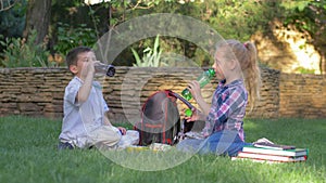 Happy young friends together drink juice from bottles sitting near backpacks on lawn during lunch in schoolyard