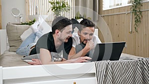 Happy young Gay couple of men are using modern laptop computer lying in bed in the bedroom. Cute male gay couple