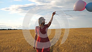 Happy young girl with brown hair walking through golden wheat field and holding balloons in hand. Beautiful woman in red
