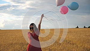 Happy young girl with brown hair walking through golden wheat field and holding balloons in hand. Beautiful woman in red