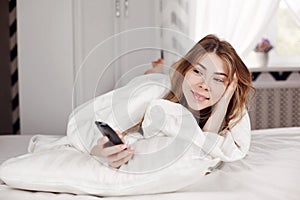 A happy young girl covered with a blanket is lying on the bed, holding the remote control from the TV