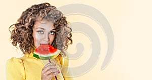 Happy young girl with curly hair holding slice of a watermelon at her face isolated over yellow background. Afro girl holds