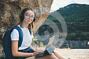 Happy young girl with glasses and backpack enjoying summer holidays listen music in headphones and laptop in nature, woman