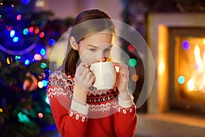 Happy young girl having a cup of hot chocolate by a fireplace in a cozy dark living room on Christmas eve. Celebrating Xmas at