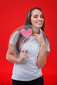 Happy young girl, having fun smiling and holding a small pink paper heart on a stick. Red background.