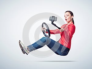 Happy young girl in jeans and red jacket driving car and making selfie with a smartphone, on light background.