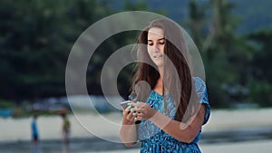 Happy young girl with long hair in a blue dress speaks on the phone on the sunny coast among palms and mountains