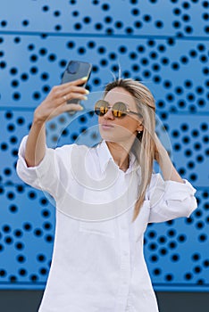 Happy young girl making funny face while taking pictures of herself through cell phone, over blue background