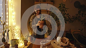 A happy young girl stands in the middle her home greenhouse and holding a clay pot with a houseplant in her hands. Slow
