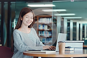 Happy young girl student studying at the college library, sitting at the desk, using laptop computer