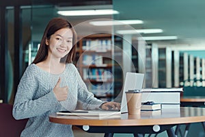 Happy young girl student studying at the college library, sitting at the desk, using laptop computer