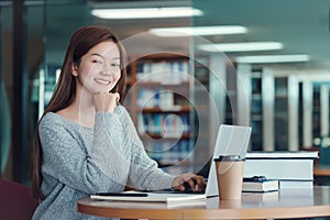 Happy young girl student studying at the college library, sitting at the desk, using laptop computer