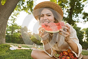 Happy young girl in summer hat having a picnic at the park, sitting on a grass