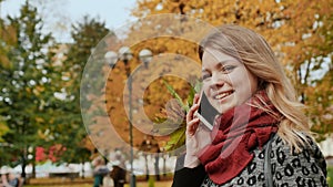 A happy young girl is talking on a mobile phone in the autumn park of the city among the colorful autumn period trees.