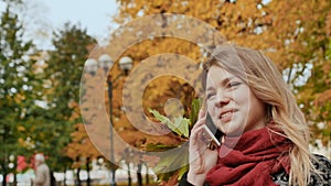A happy young girl is talking on a mobile phone in the autumn park of the city among the colorful autumn period trees.