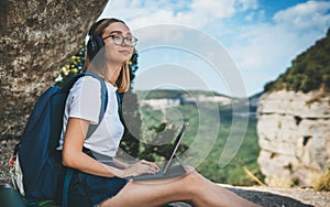 Happy young girl tourist with backpack enjoying summer holidays listen music in headphones and laptop in nature, woman copywriter