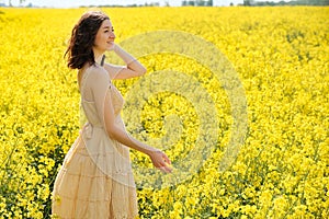 Happy young girl walking in canola field on summer day. Beautiful woman in beige dress enjoying nature