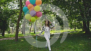 Happy young girl walks in the green sunny park and holds colourful balloons