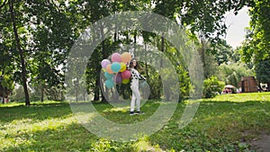 Happy young girl walks and poses with colourful balloons at camera in park.