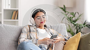 Happy young girl with white headphones chilling and listening her favorite song. Indian brunette woman seat on sofa and