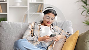 Happy young girl with white headphones chilling and listening her favorite song. Indian brunette woman seat on sofa and
