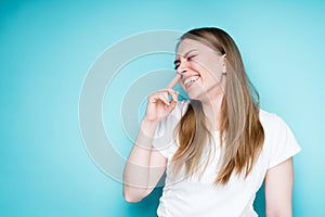 Happy young girl in a white t-shirt touches her nose with her finger and laughs while standing on a blue background