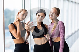 Happy young girls in sportswear taking selfie in gym. Three female friends in healthclub taking selfie with mobile phone