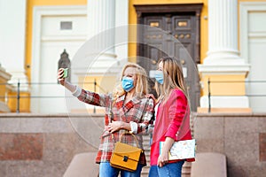 Happy young girls taking selfies in city. Two women in face masks walking at street. Friends travel at city together