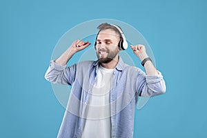 Happy young guy in headphones dancing to his favorite song, listening to music on blue studio background