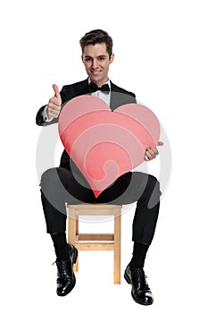 Happy young guy holding big red heart and making thumbs up sign