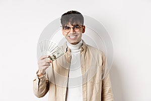 Happy young guy holding dollar bills and smiling, making money and looking cheerful at camera, standing on white