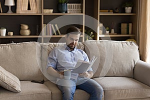 Happy young guy read good book drinking tea on sofa