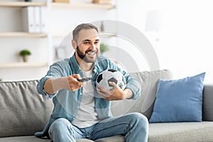 Happy young guy watching soccer game on TV, holding remote control and ball at home