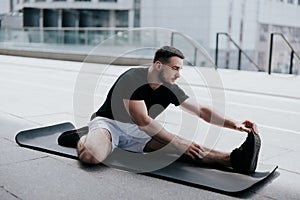 Happy young handsome brunette man in sportswear warming up before street workout stretching on yoga mat, healthy fit