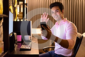 Happy young handsome Indian businessman video calling with phone while working overtime at home