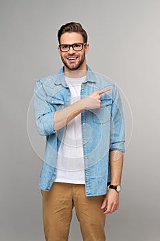 Happy young handsome man in jeans shirt pointing away standing against grey background