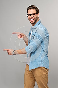 Happy young handsome man in jeans shirt pointing away standing against grey background