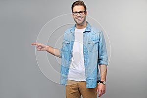 Happy young handsome man in jeans shirt pointing away standing against grey background